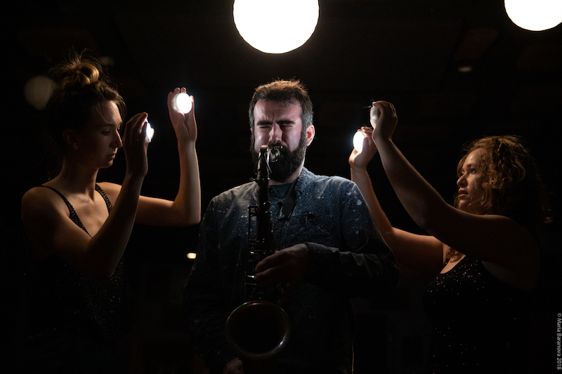 Two women hold lights while one man blows into a saxophone